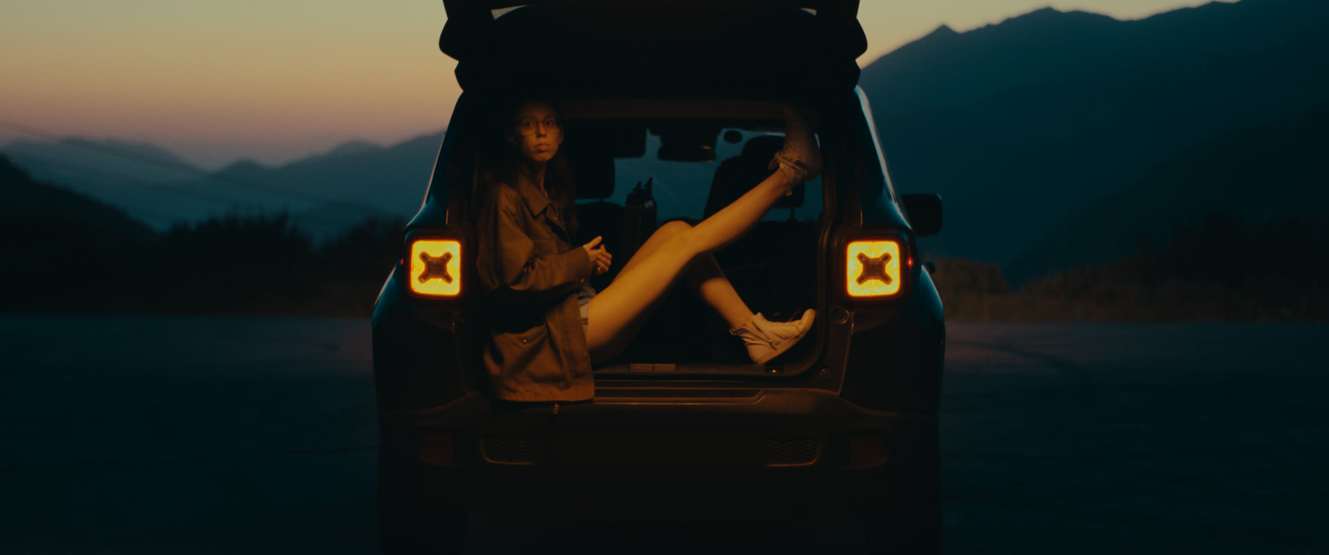 A woman sits in the open car trunk at dusk, taillights glowing — still from Memories